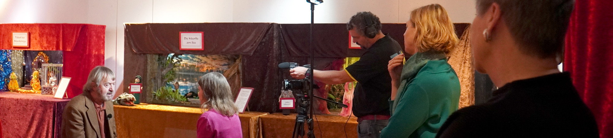 In der Bildmitte Baut der Kameramann seine Technik auf. Auf der linken Seite sind Kurator und Redakteurin im Gespräch. Zwei Zuschauer beobachten die Szene.